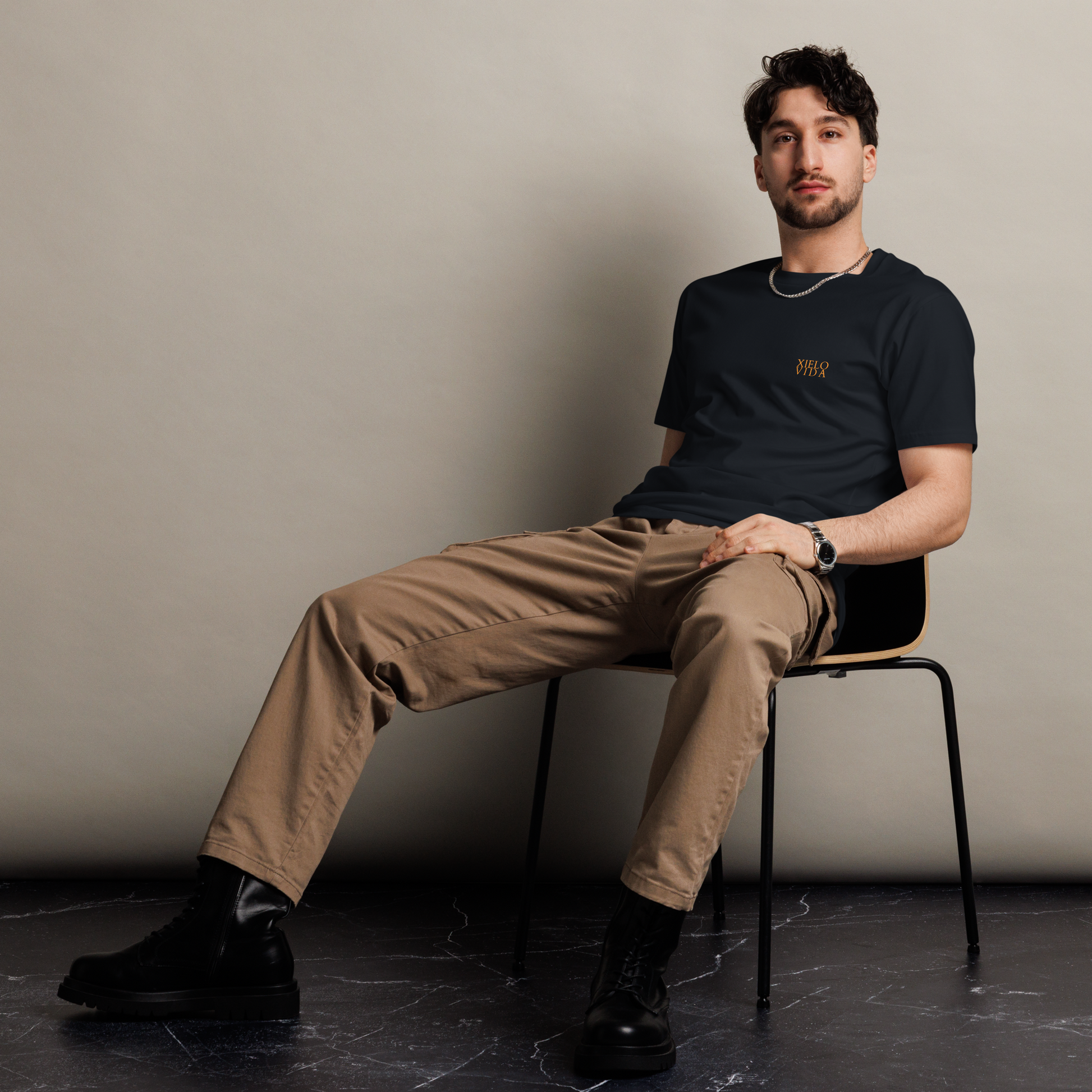 Male model seated in a relaxed pose wearing a navy premium mens t-shirt with the Xielo Vida gold logo on the left chest, paired with khaki pants and black combat boots.